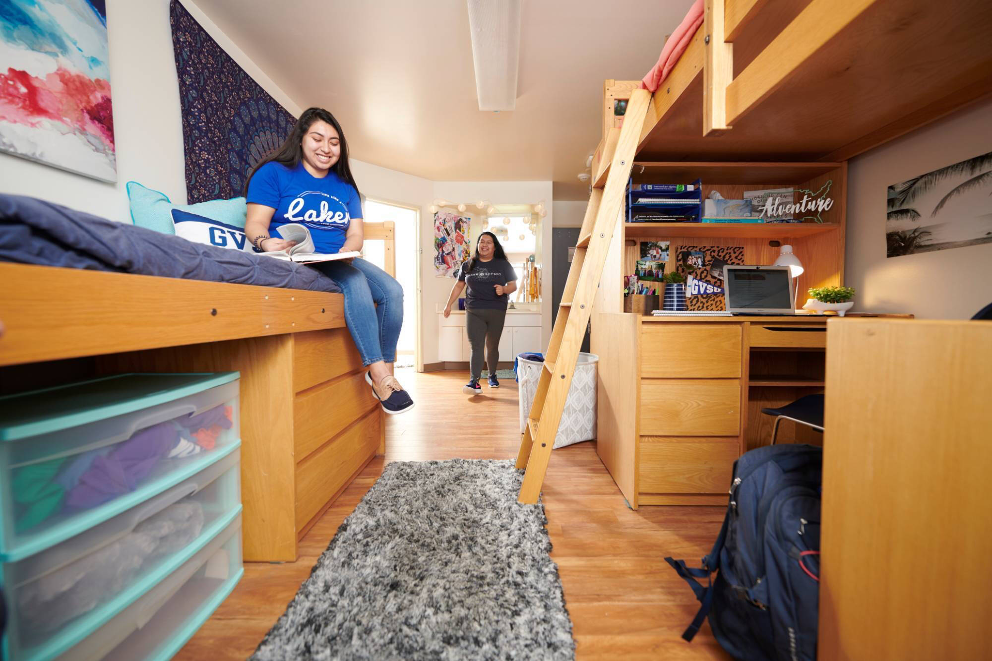 Two young women are in a cozy, well-organized suite style bedroom. One sits on a lofted bed reading, the other enters smiling. The room has warm wood furniture.